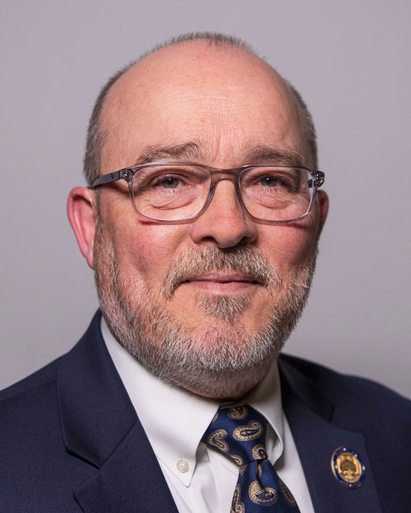 Portrait of a person with glasses, wearing a suit and tie, with a closely cropped beard and a gentle expression.