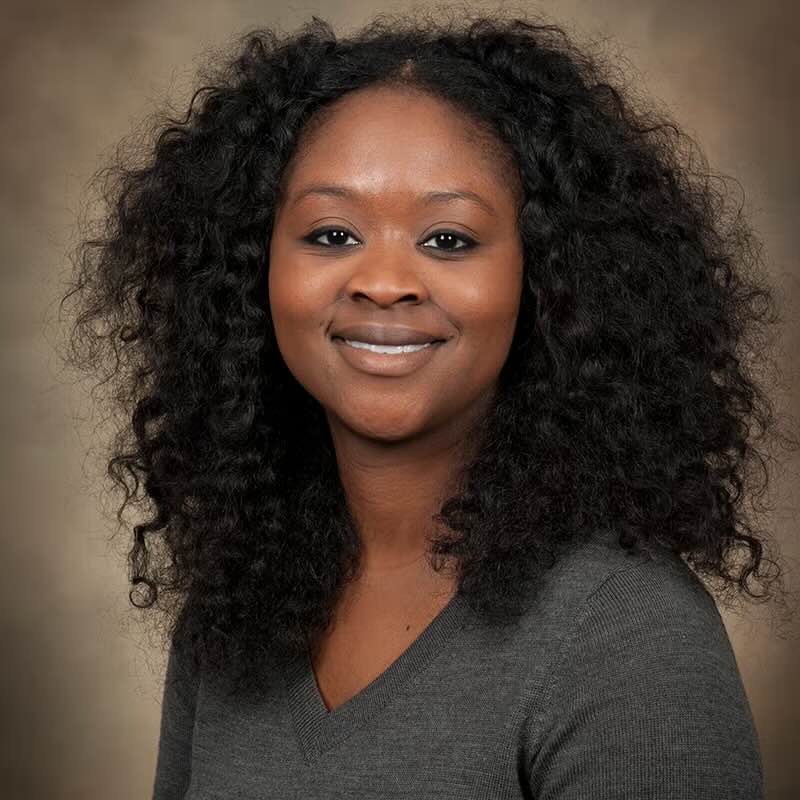 Close-up of an individual with curly black hair smiling in front of a beige background.