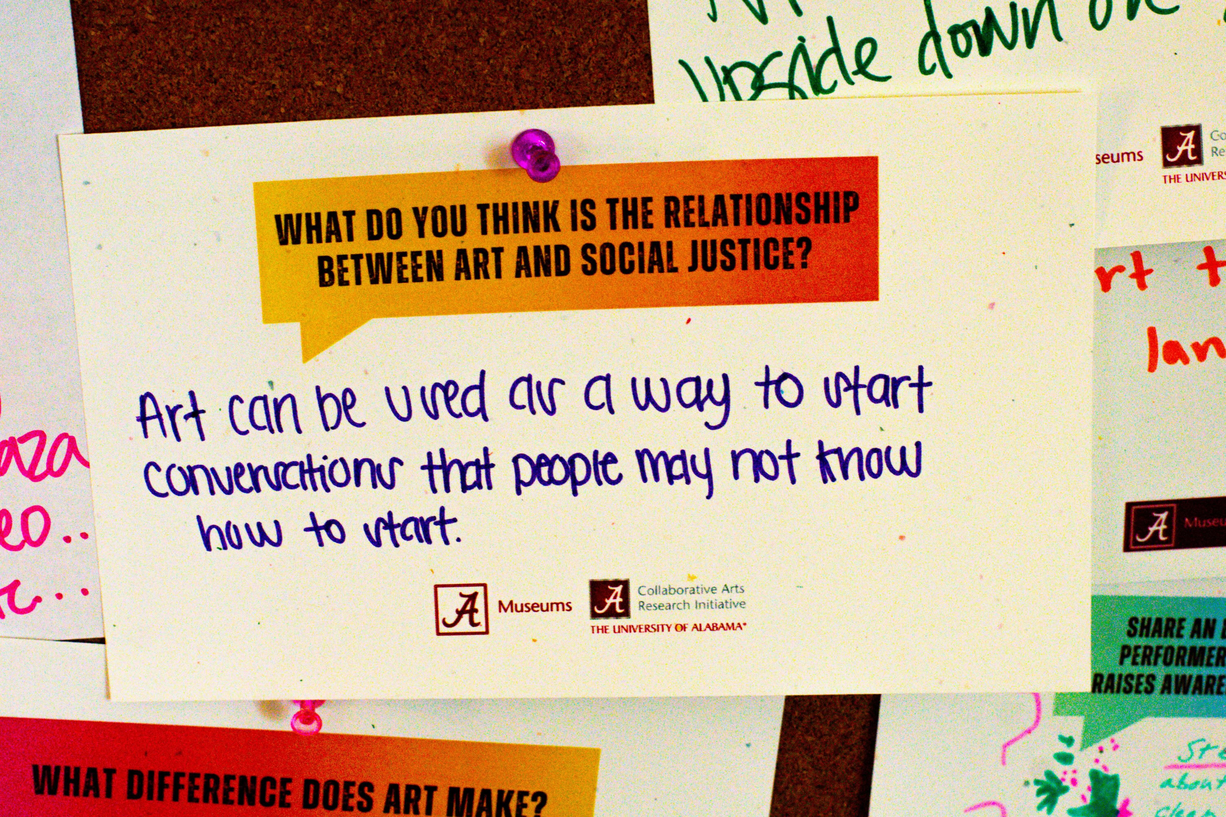 Bulletin board featuring a large yellow note with a question about the relationship between art and social justice, and a response on an orange note stating art's potential to start conversations. The board includes various other notes and the logos of the Collaborative Arts Research Initiative at The University of Alabama and The University of Alabama Museums.