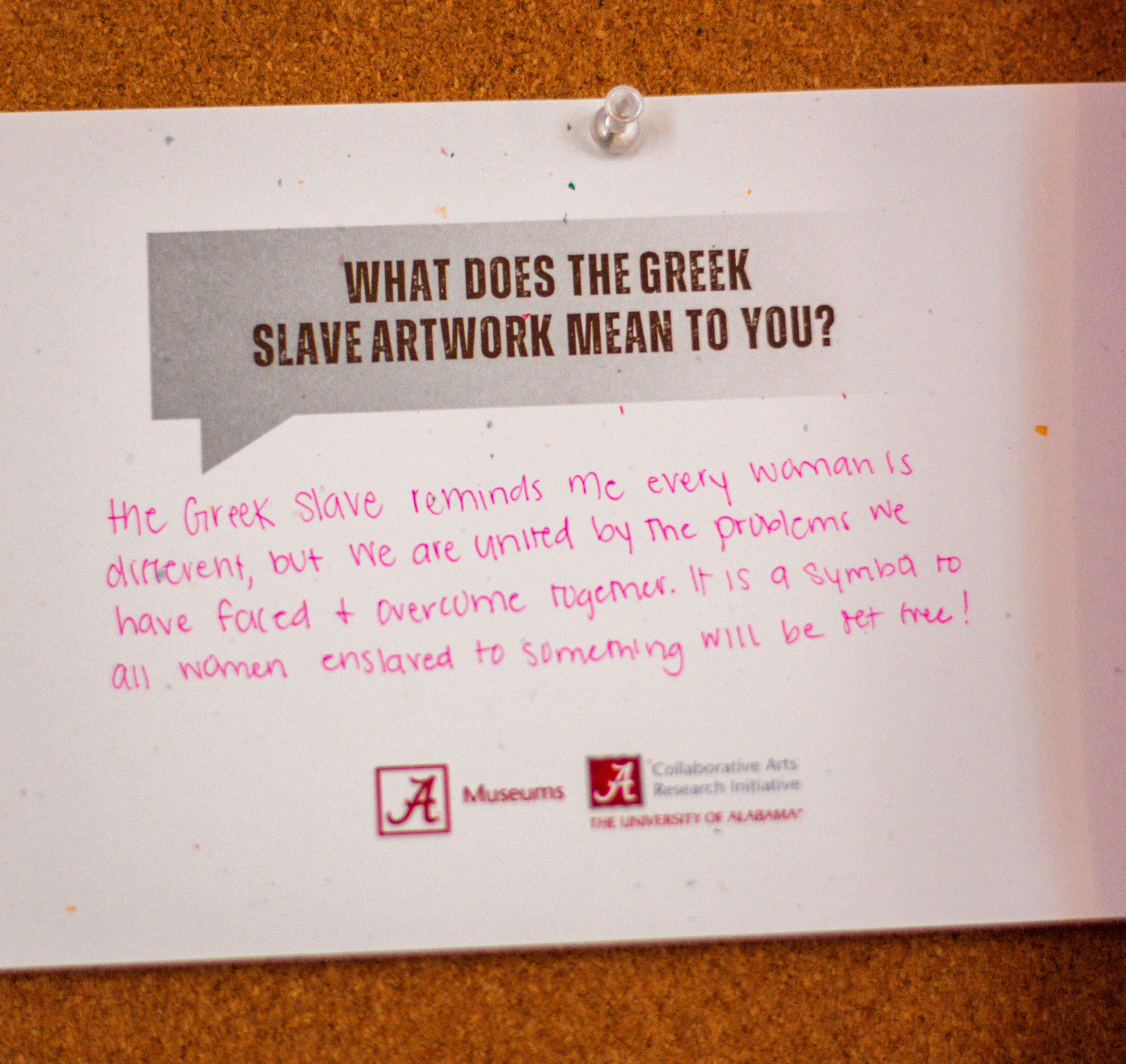 A close-up photo of a written note on an exhibit feedback wall asking, "What does the Greek Slave artwork mean to you?" The response mentions it as a symbol of universal struggles against stereotypes and oppression. The logos of The University of Alabama Museums and Collaborative Arts Research Initiative are visible at the bottom.