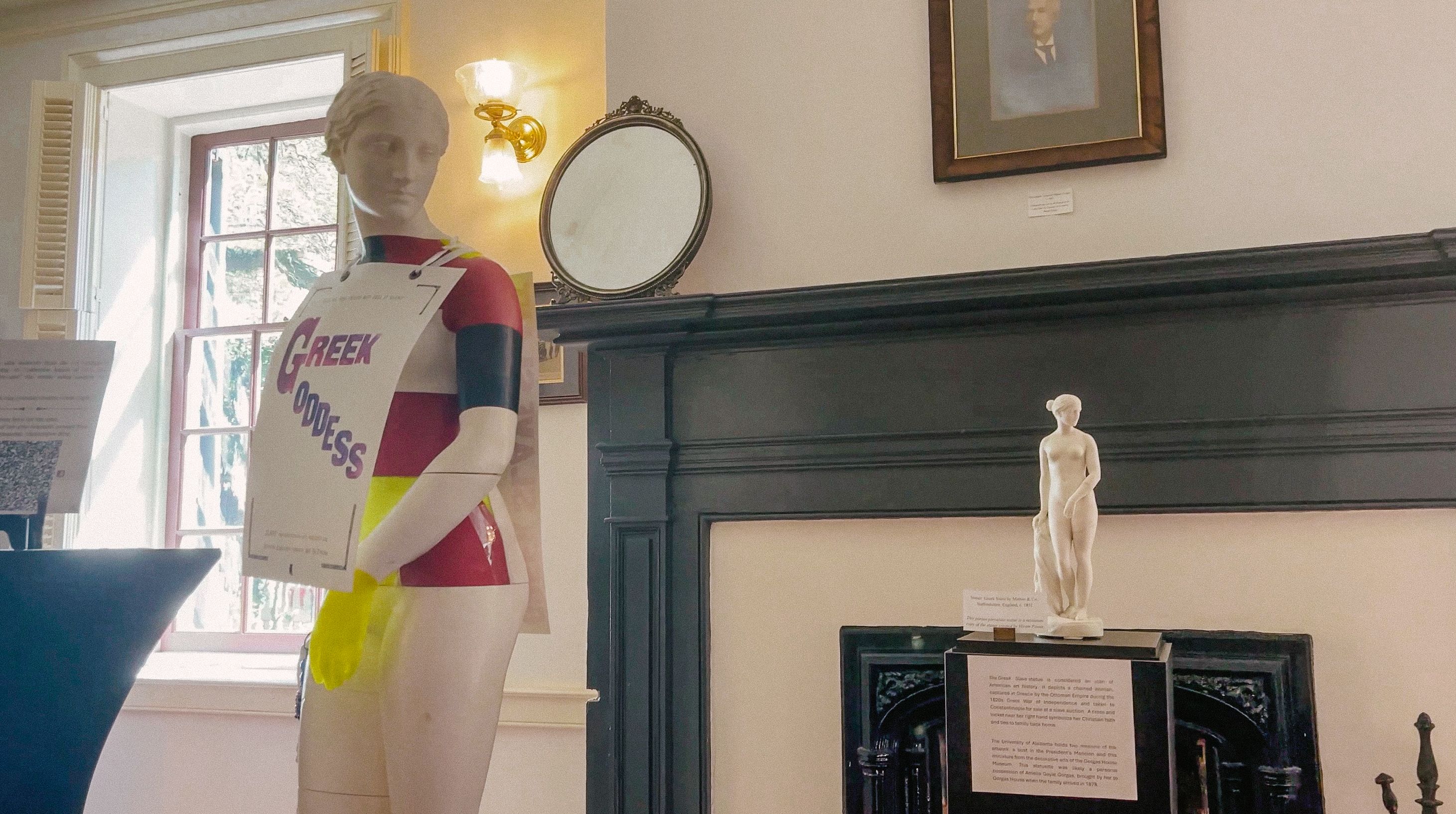 A mannequin dressed in a colorful top with a sign that reads "Greek Goddess" hanging on it and displayed in a room with classical decor, featuring a small statue, ornate mirror, and framed paintings.