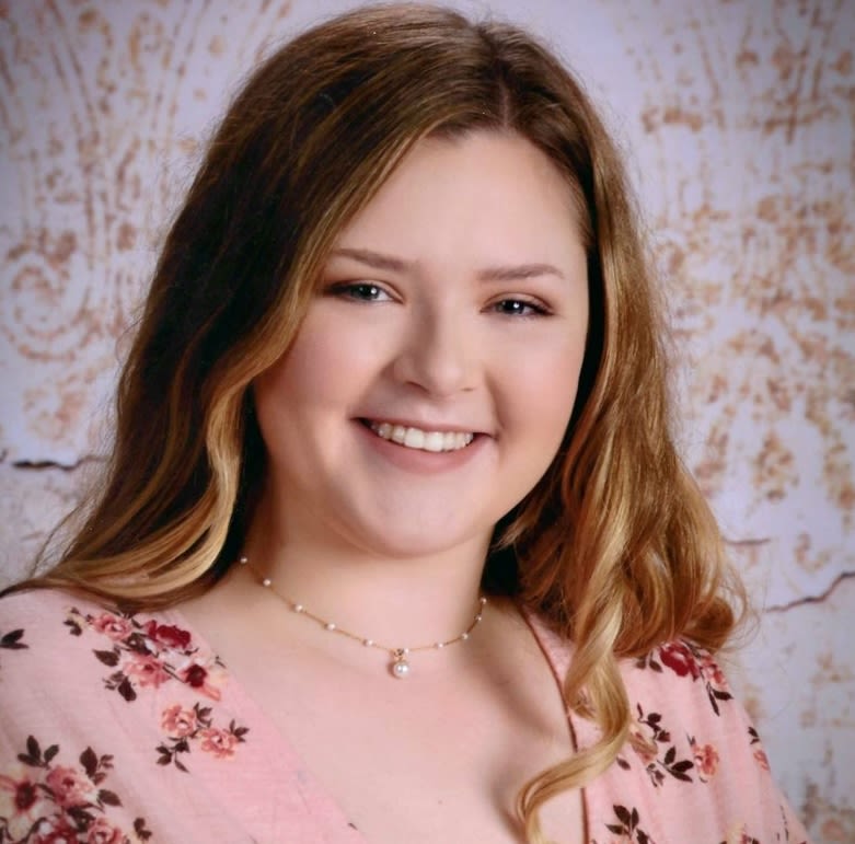 Portrait of a smiling individual with shoulder-length hair, wearing a floral top and a necklace.