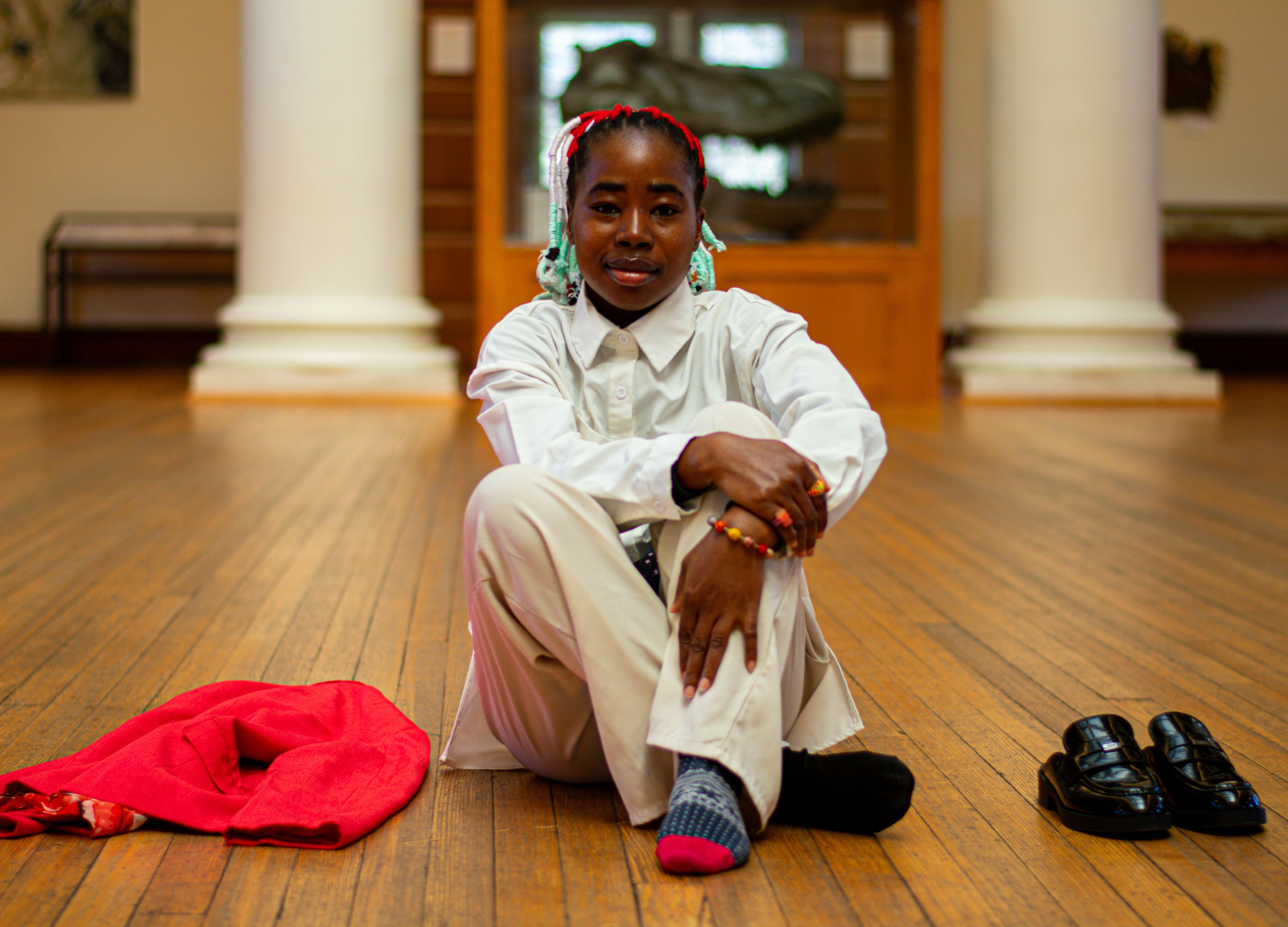 Individual sitting on the floor with colorful braids, wearing a white shirt and adorned with bracelets, next to a red scarf and black shoes.