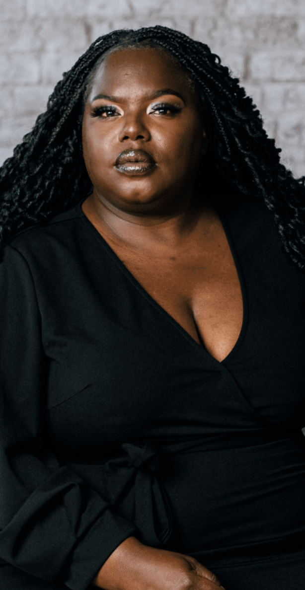 Portrait of a person with long braided hair, wearing a black dress, seated against a brick wall background.