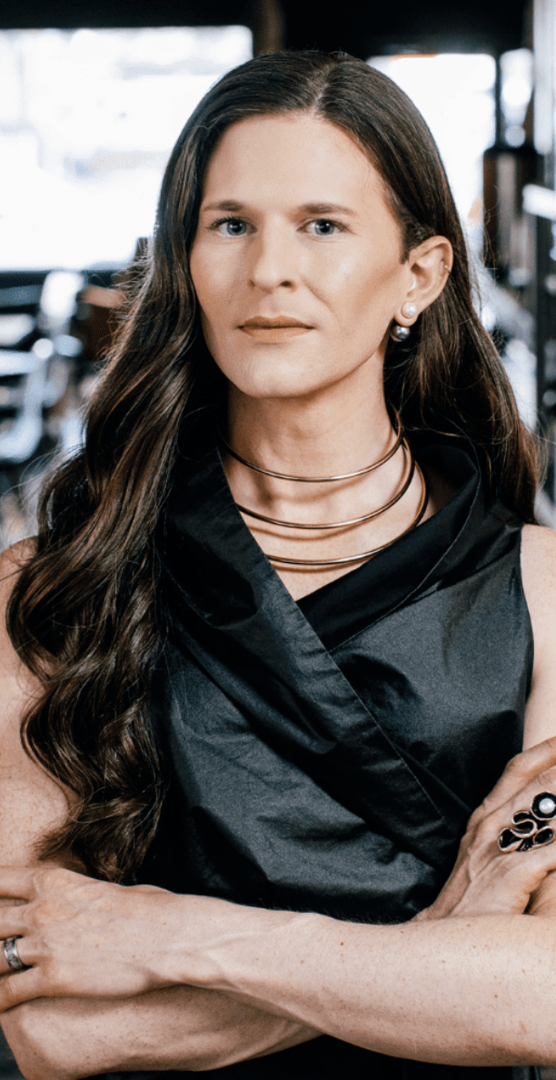 Portrait of a person with long, straight hair wearing a black sleeveless top and layered necklaces, sitting confidently in an industrial setting.