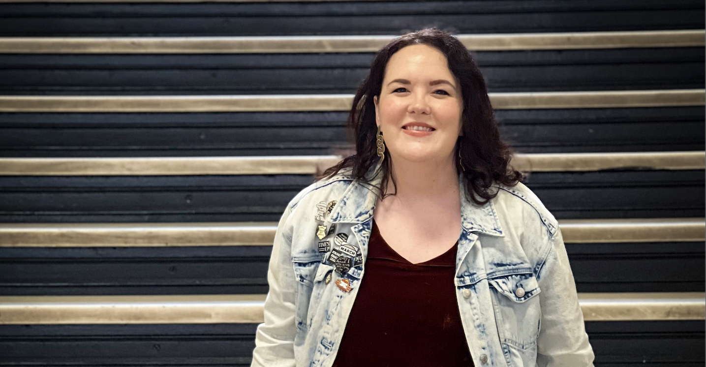 Person smiling on black stair steps, wearing a denim jacket over a red top.