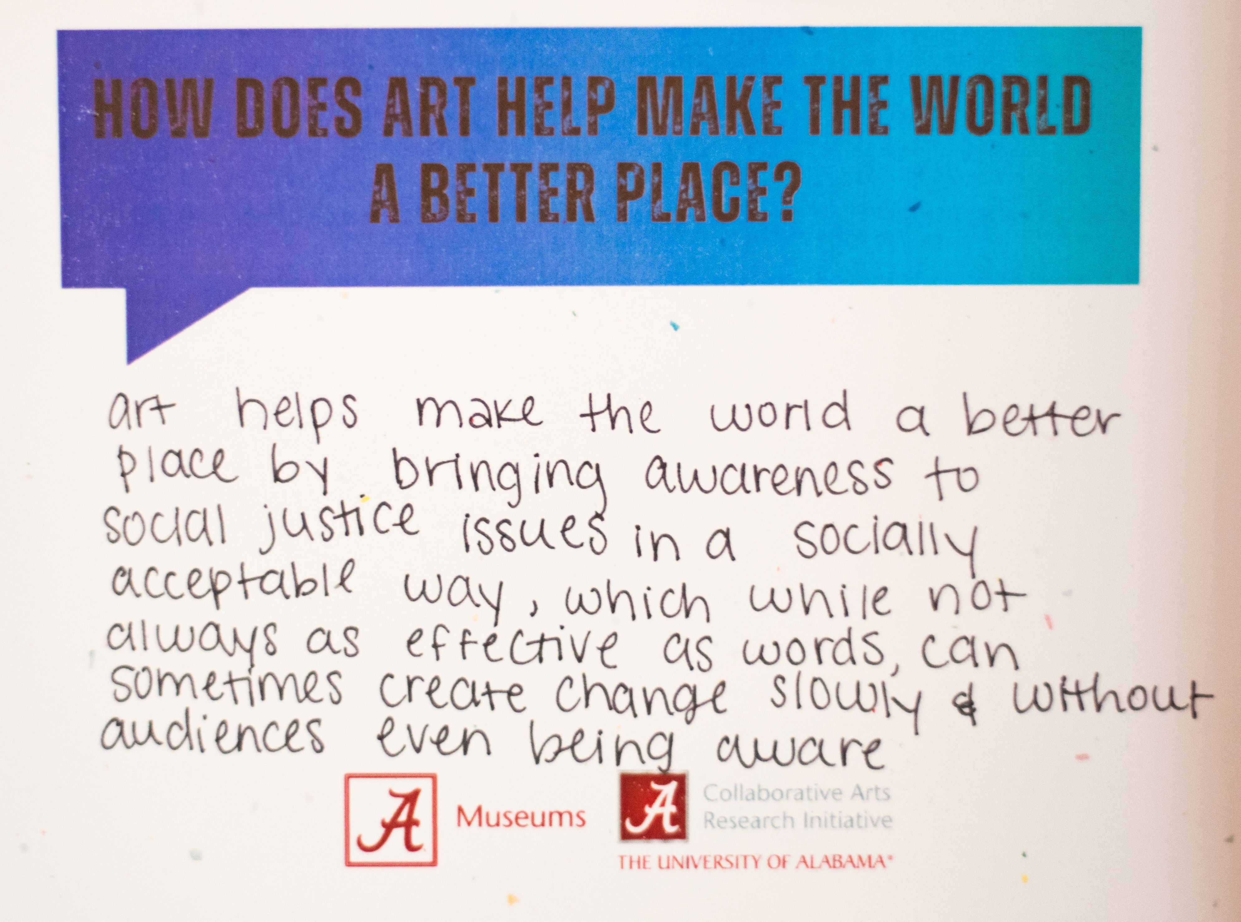 Text on a presentation board asking "How does art help make the world a better place?" with a handwritten response about art raising awareness and sparking change, including logos for The University of Alabama Museums and The University of Alabama's Collaborative Arts Research Initiative.