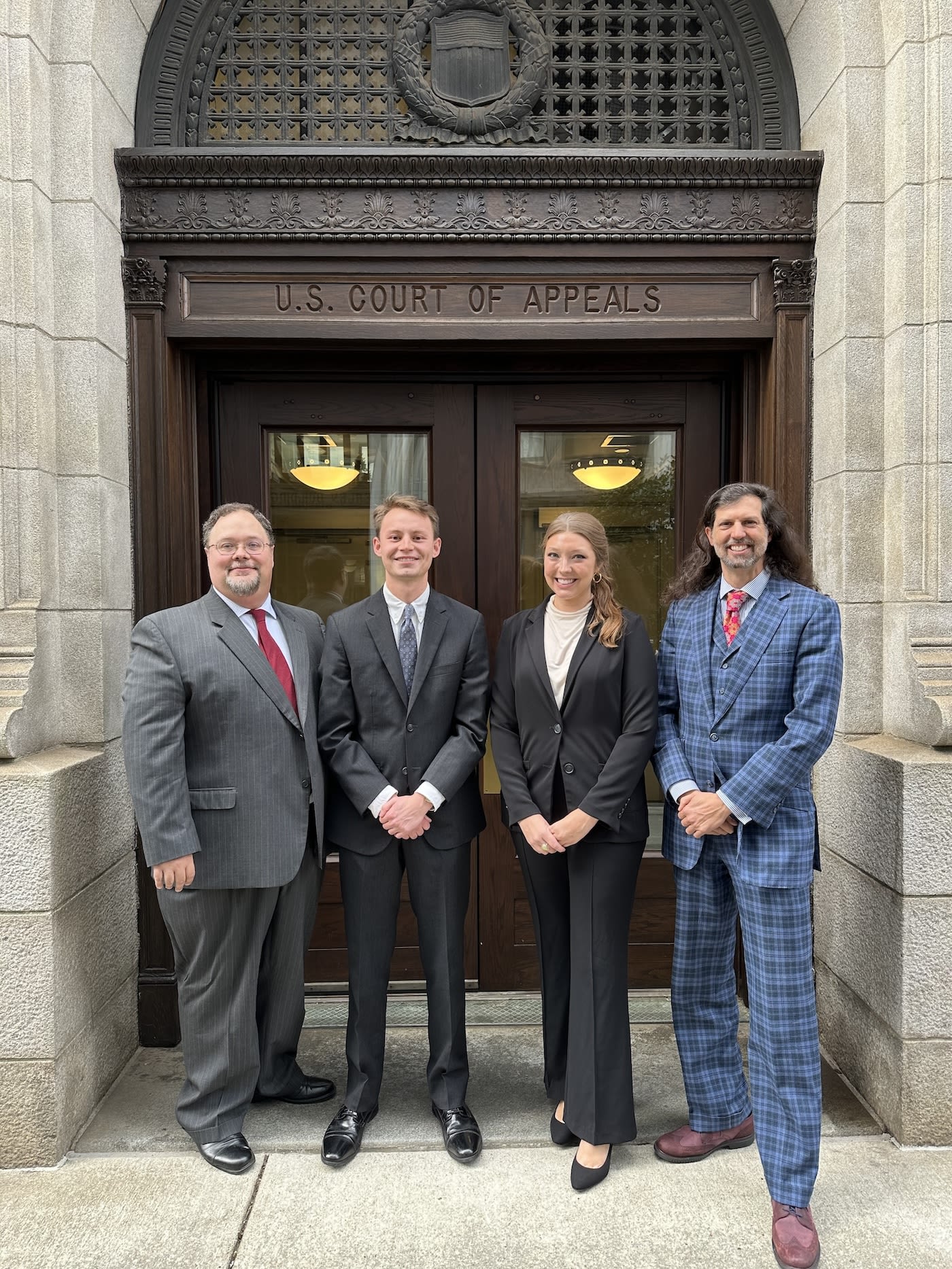 Four people dressed in suits stand in front of a courthouse building