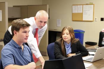 A man in a button down shirt with a red tie leans over the computer of a male student, pointing at the screen, with a female student looking on. 