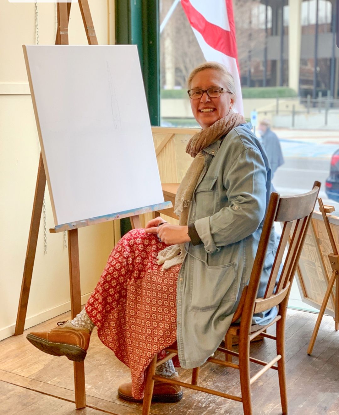 Photo of a woman, Sonya Clemons, seated in a wooden chair in front of a blank canvas sitting on an easel. She is wearing a red dress with a white pattern, a coat, and a scarf.
