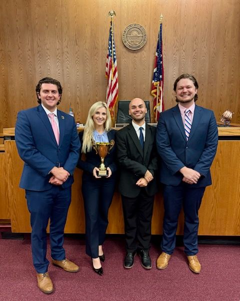 Four law students dressed in business attire stand at the front of a courtroom. One students is holding a trophy.