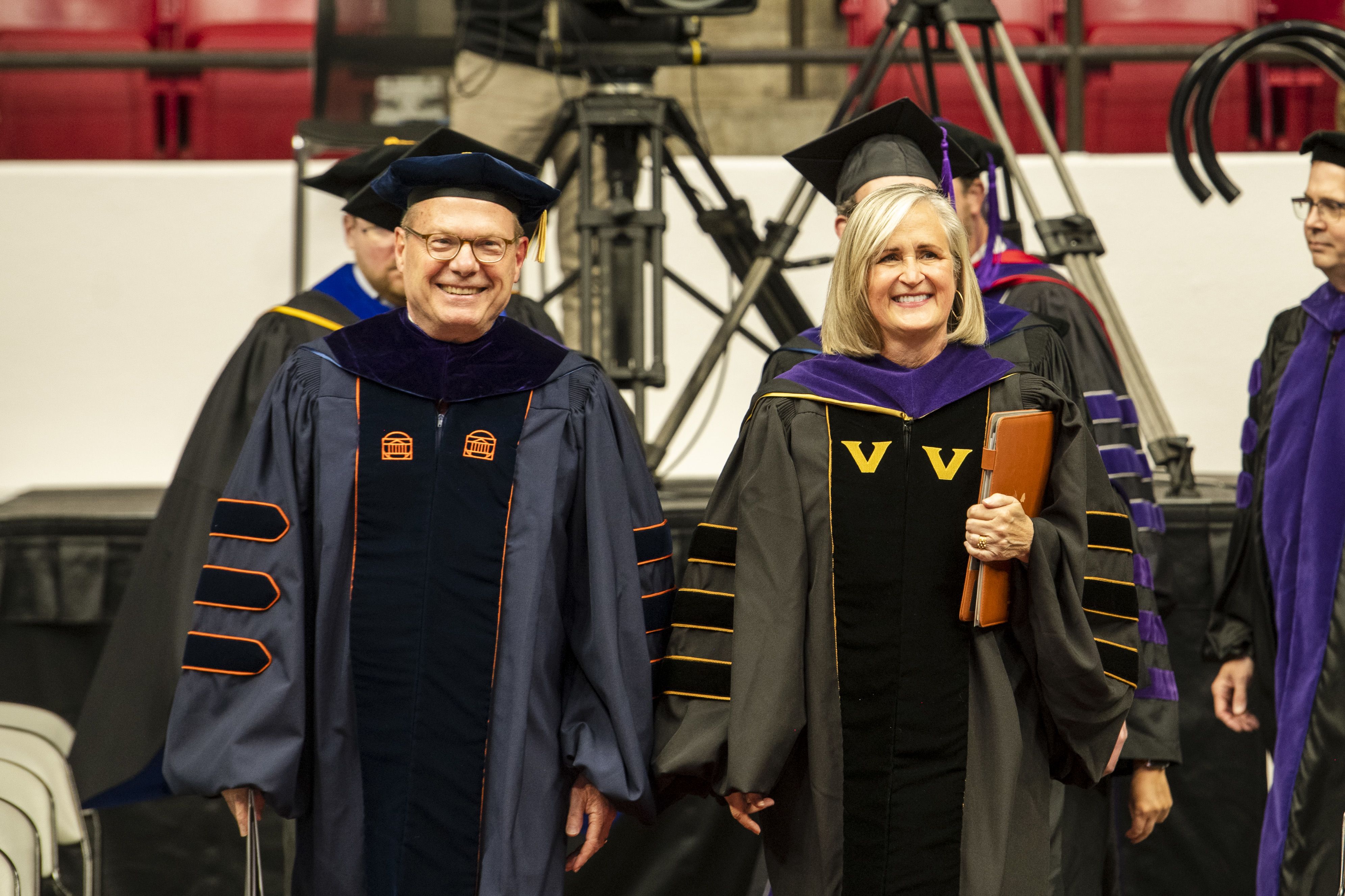 Two people dressed in graduate robes smile while walking