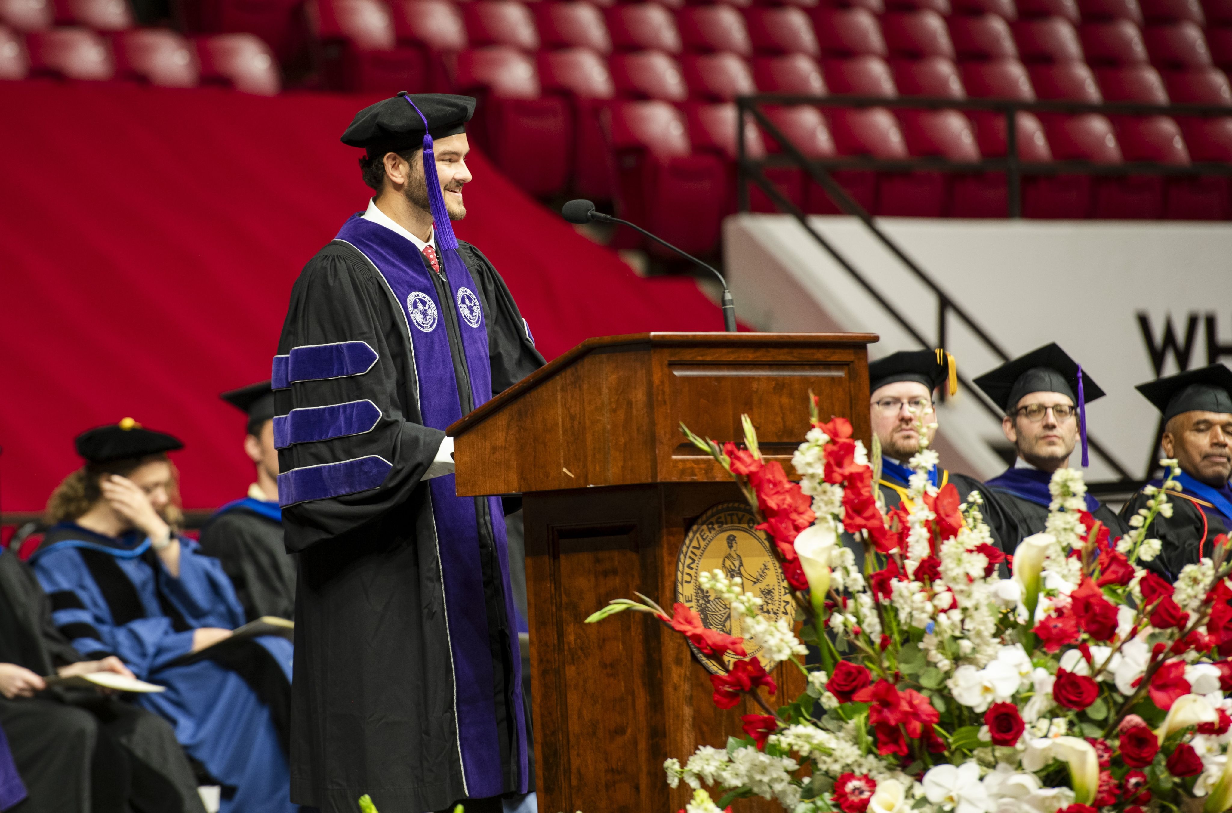 A young man wearing a graduate cap and robes speaks from behind a podium.