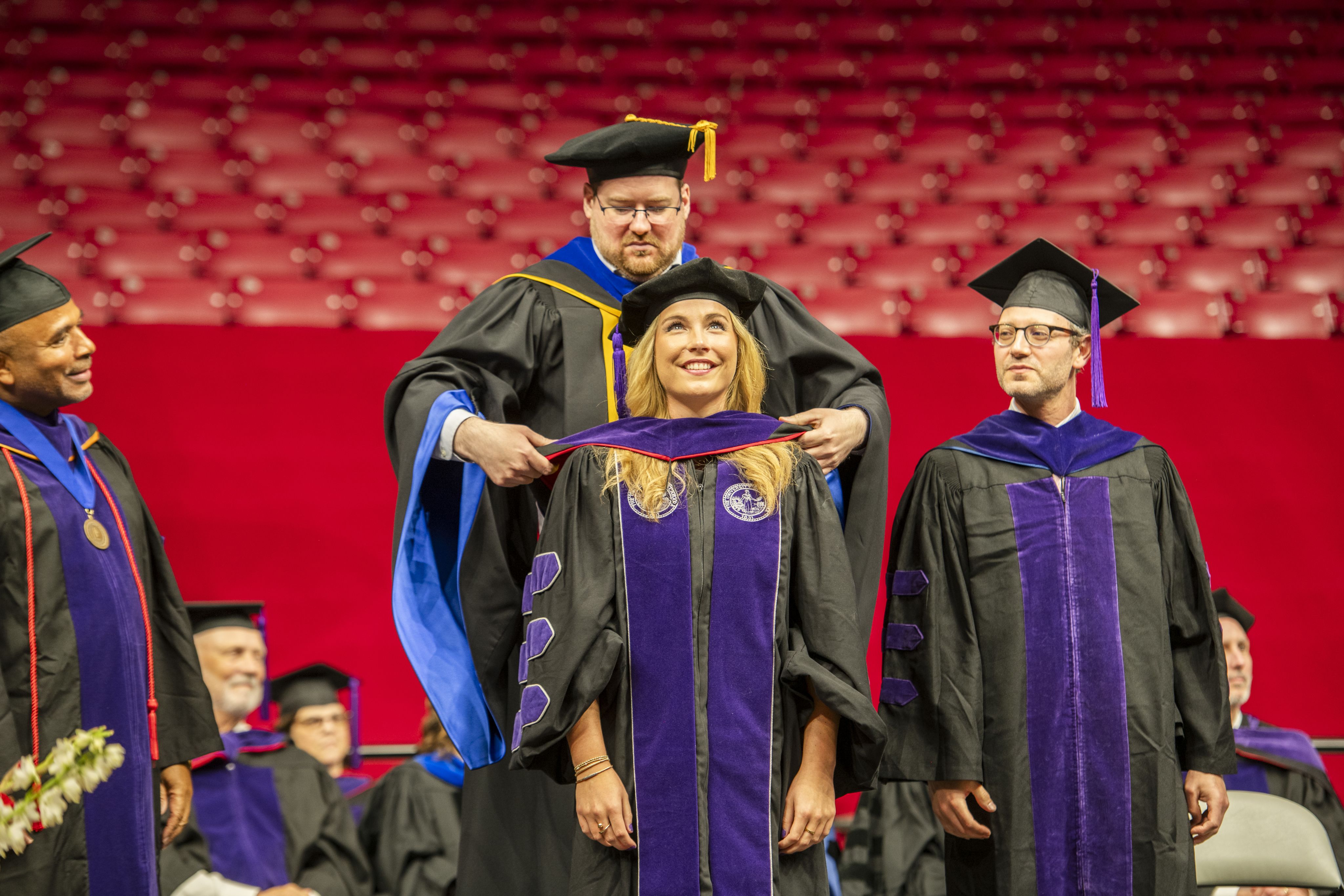 A law professor, Ben McMichael, places a hood on a law school graduate, Chrissy Tubbs, during the commencement ceremony as Professor Russell Gold looks on, smiling.