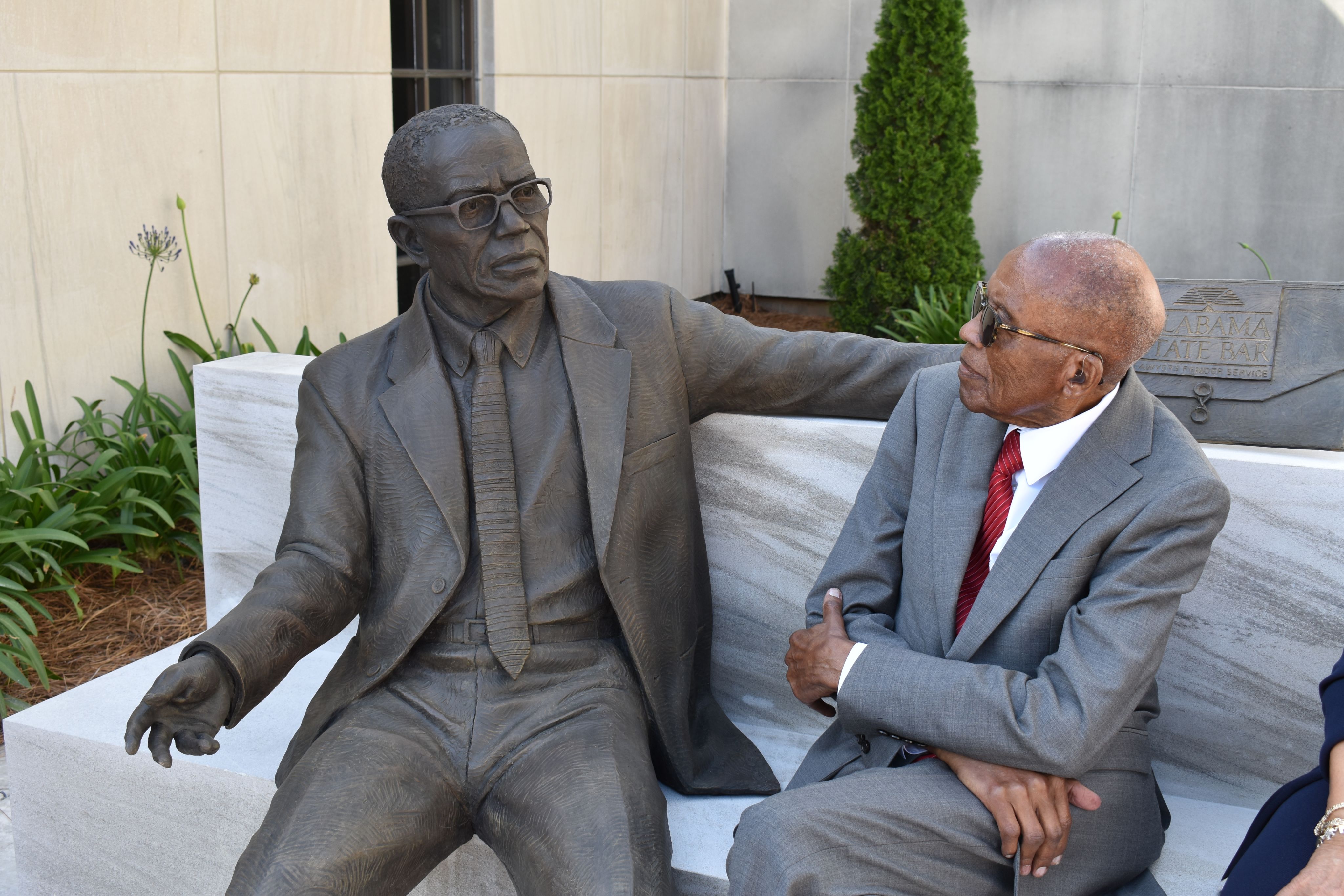 An older man dressed in a gray suit with a red tie sits on a bench looking at a statue of himself.