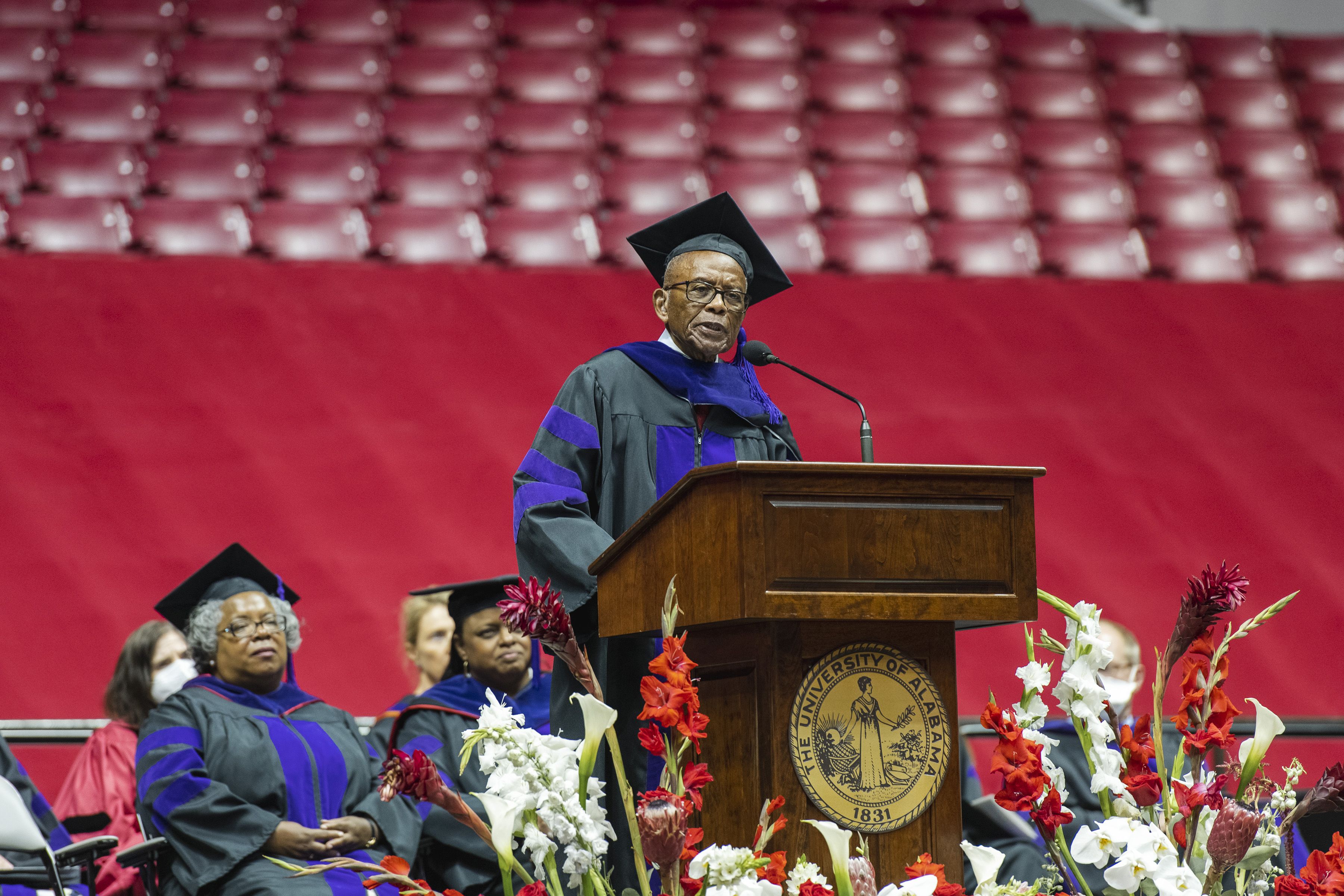 An older man dressed in a graduate cap and gown speaks from behind a podium at a graduation ceremony.