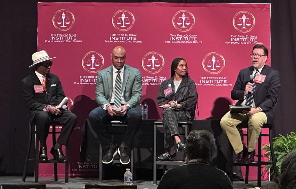 Four people–three men and one woman–sit on a stage. The man on the far right is speaking into a microphone. The background is red with a repeated logo that reads "The Fred D. Gray Institute for Human and Civil Rights."