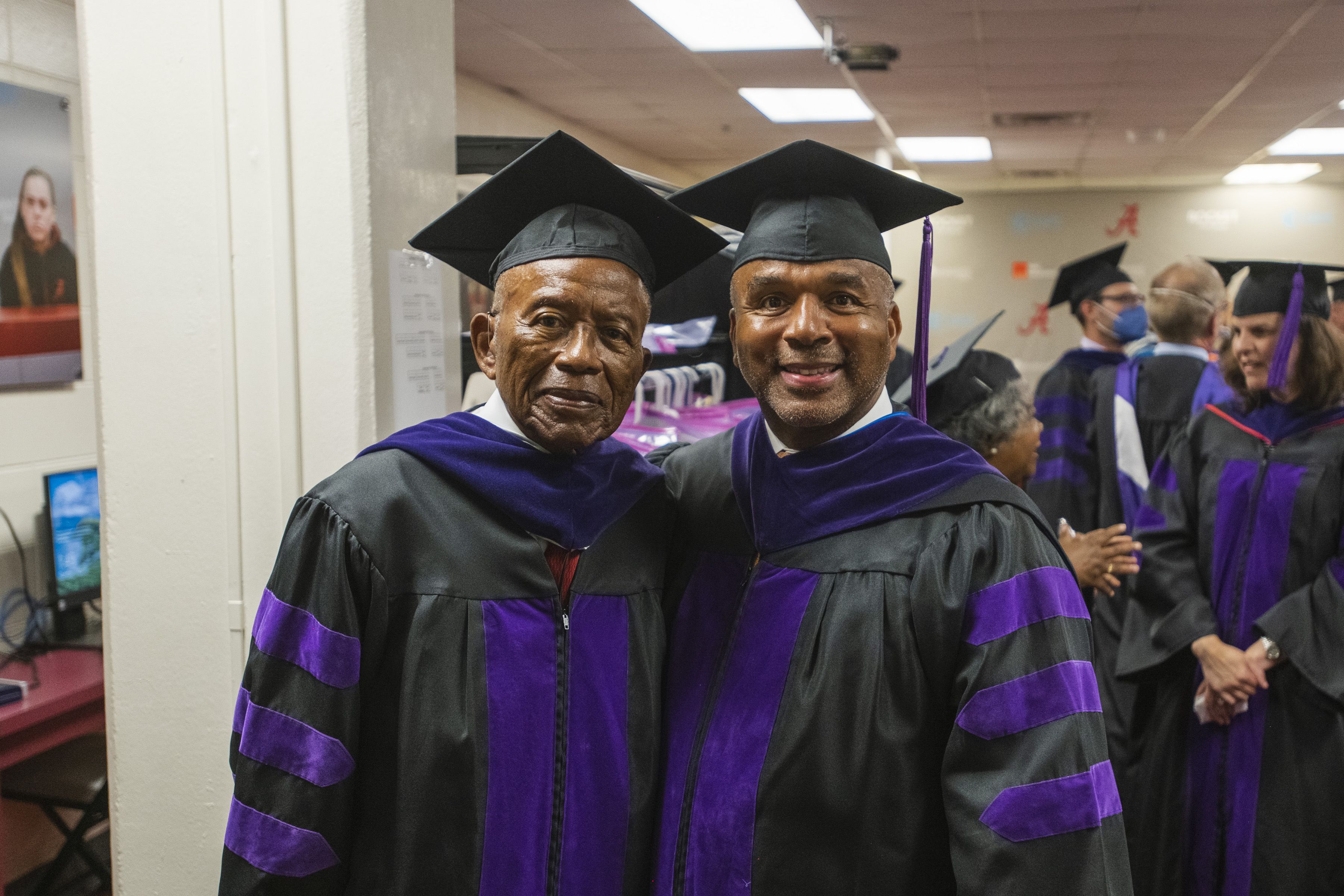 Two black men dressed in graduate caps and gowns smile at the camera before a graduation ceremony. 