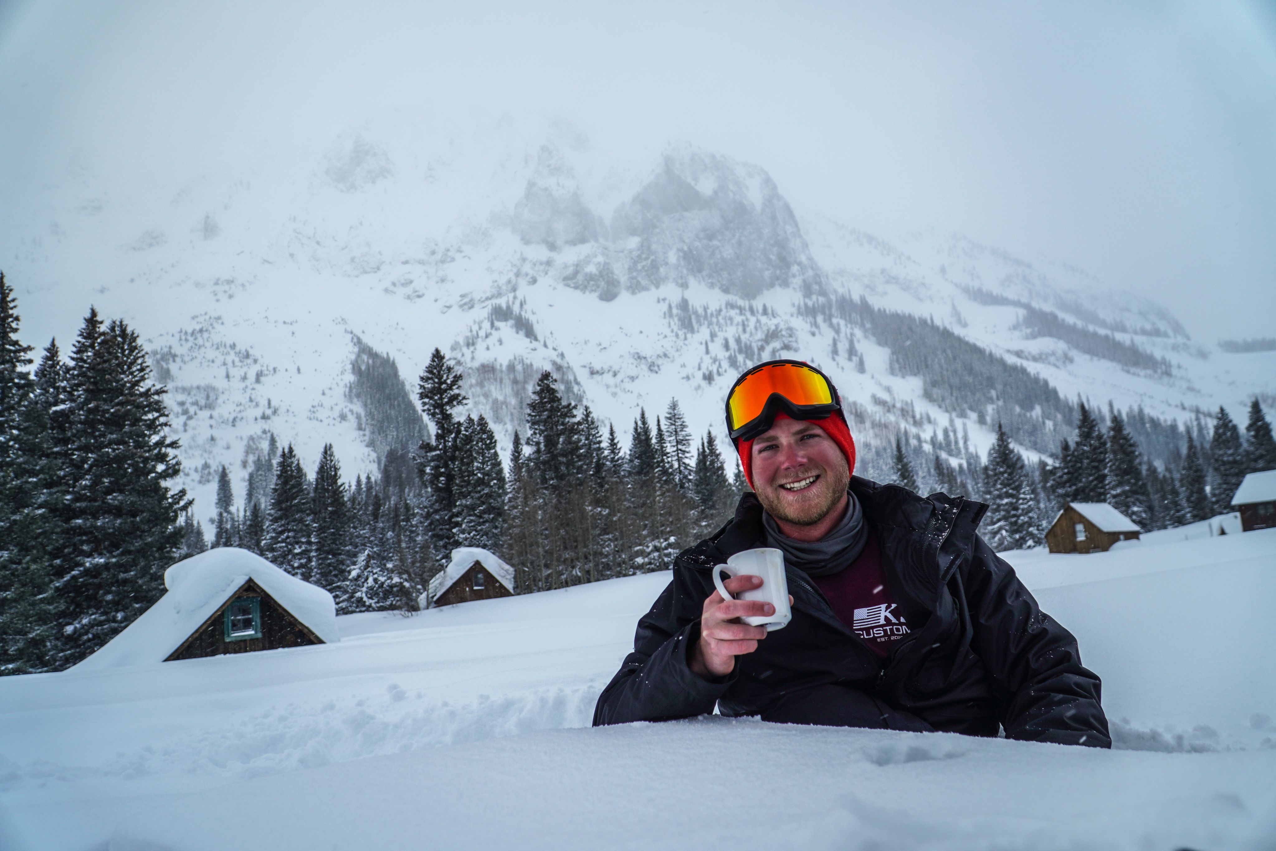 Zac smiles and holds a cup of coffee in front of snow covered cabins and mountains.