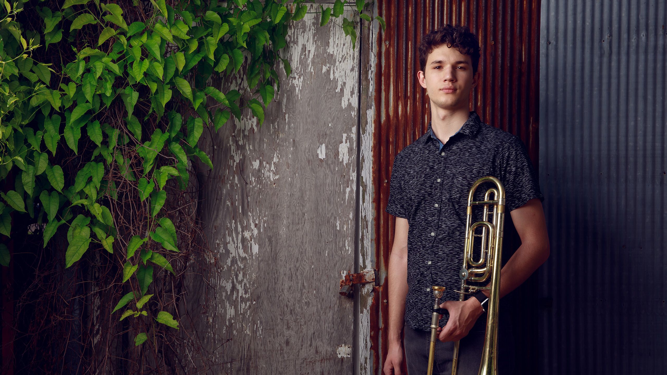 Isaiah stands holding his trombone in front of a painted shed door with a wall plant beside him.