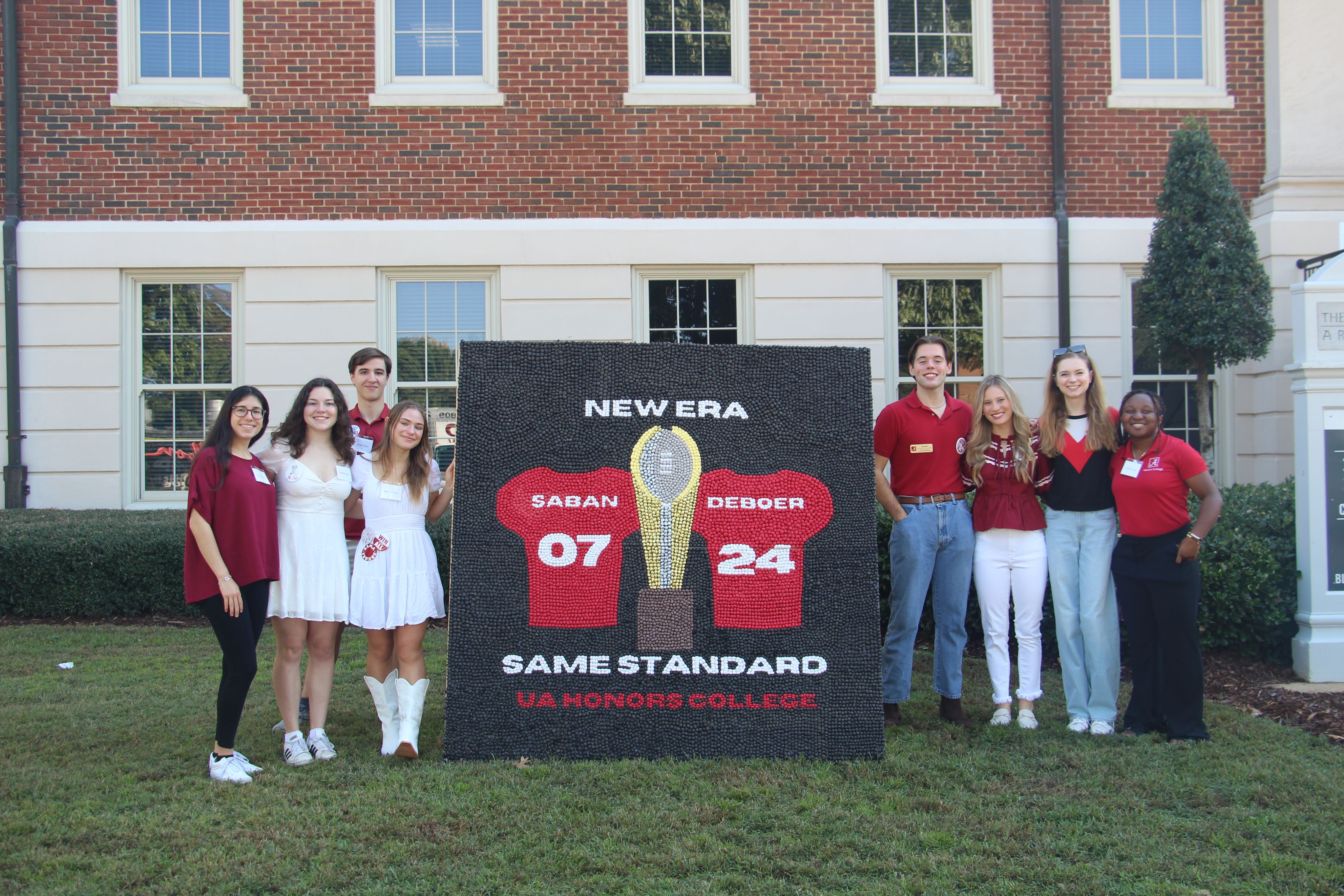 Honors students smile around their pomping project for homecoming. The pomping project reads, "New Era, Same Standard."