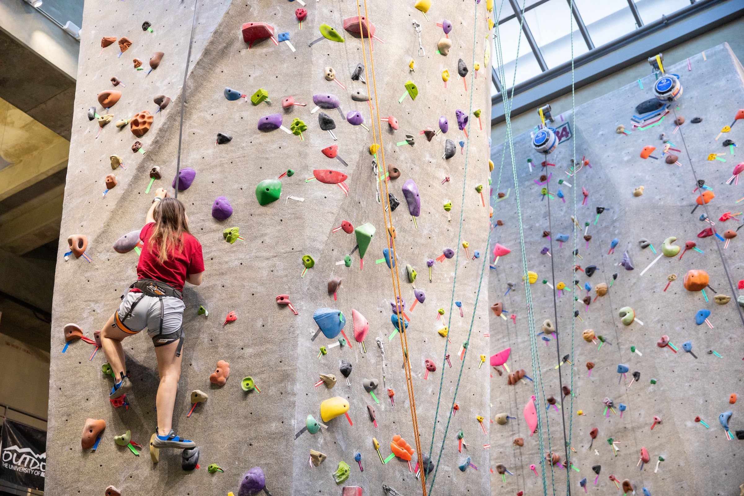 A student wearing shorts and a t-shirt is climbing the climbing wall inside the Rec Center.